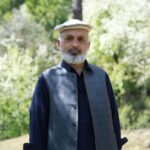 Elderly man wearing a Chitrali cap poses outdoors in a scenic natural setting.