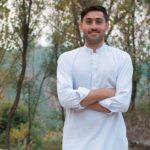 Smiling man in traditional clothes poses confidently in a serene outdoor setting.